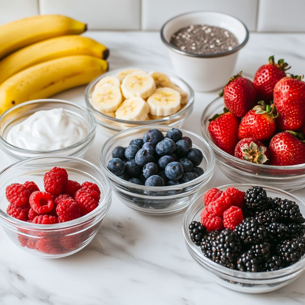 The ingredients for a berry smoothie laid out on a clean white marble kitchen counter in glass bowls.