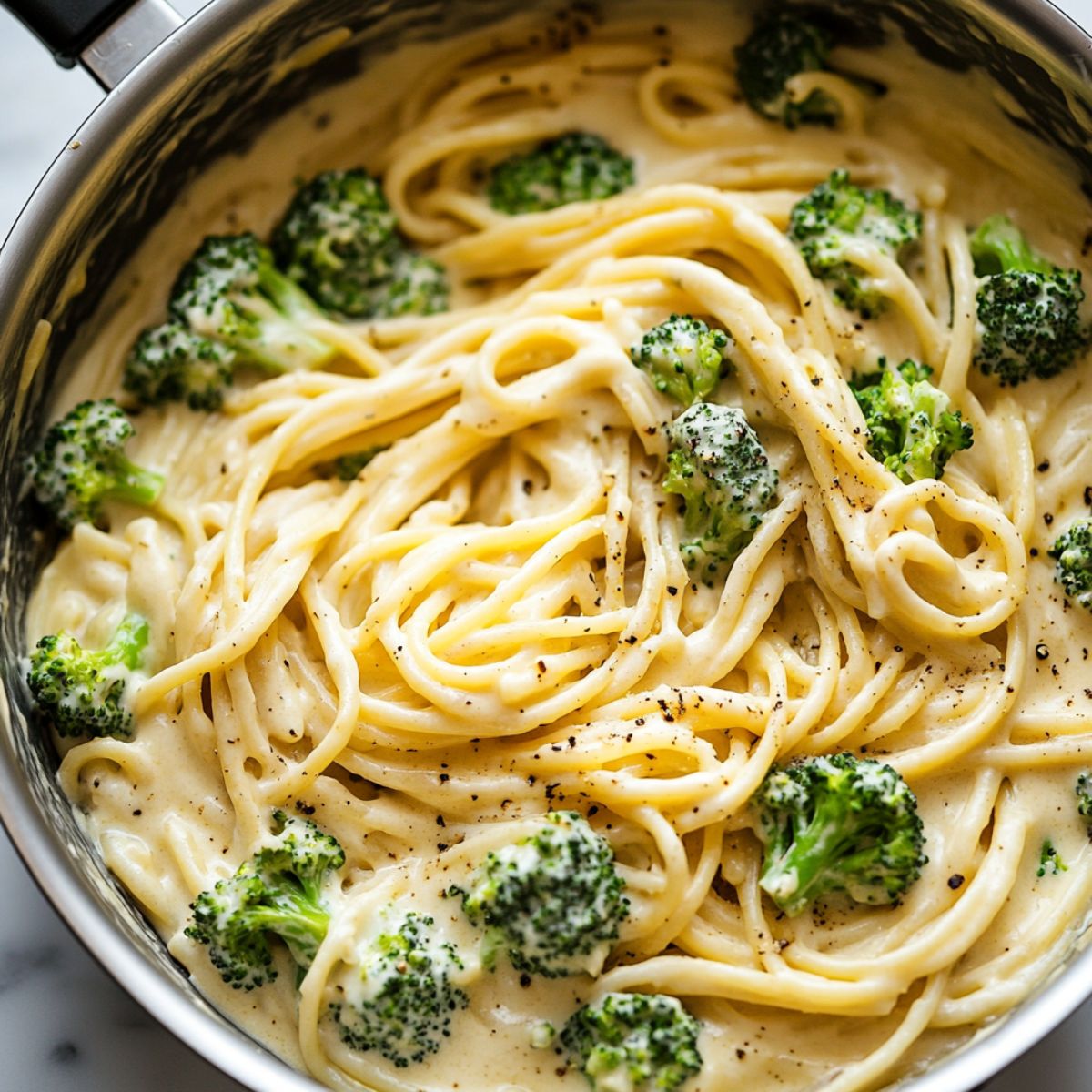 Creamy broccoli pasta being mixed in a pan on a white marble kitchen counter.