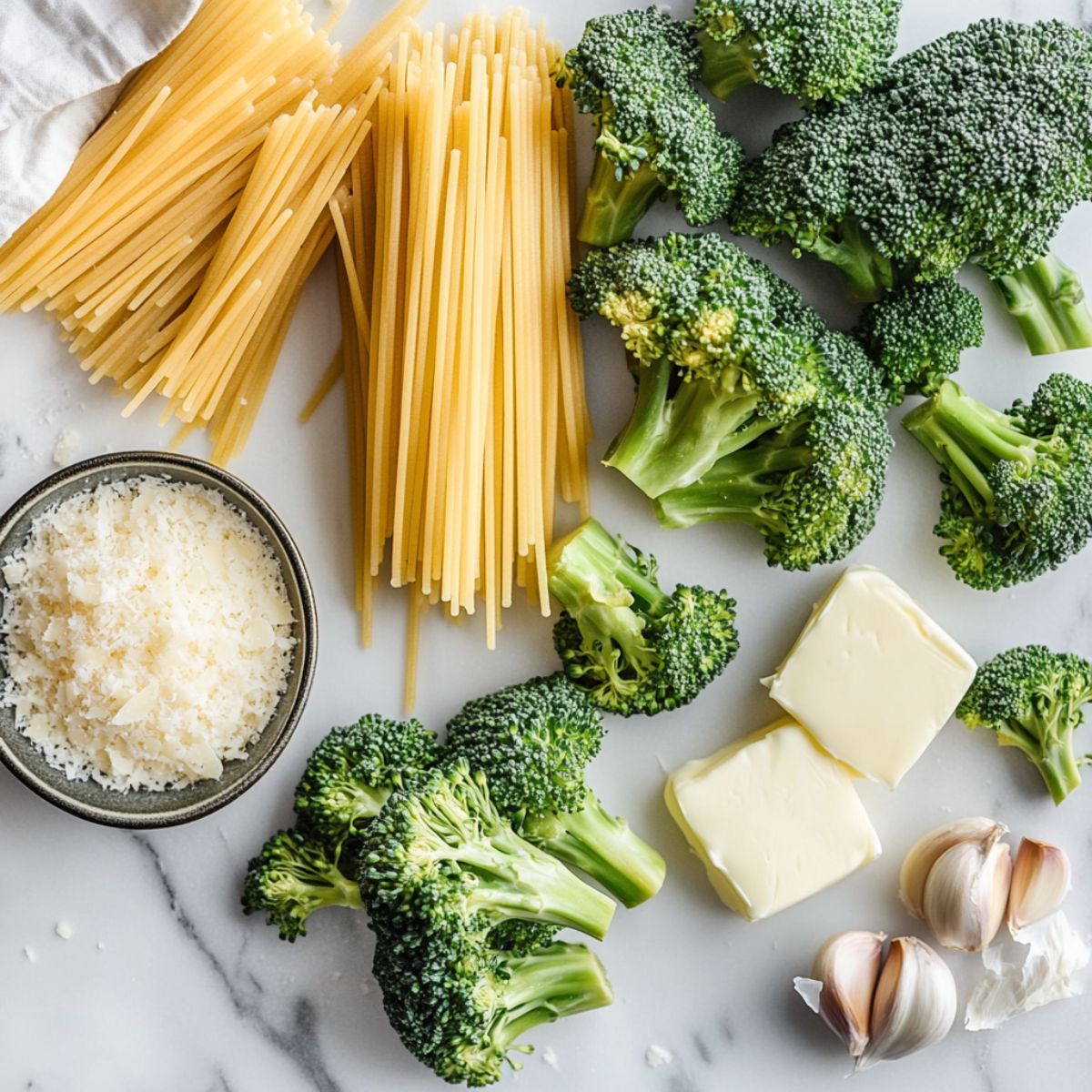 Close-up of ingredients for creamy broccoli pasta recipe including broccoli, penne, garlic, cheese, and heavy cream, on a white marble kitchen counter.
