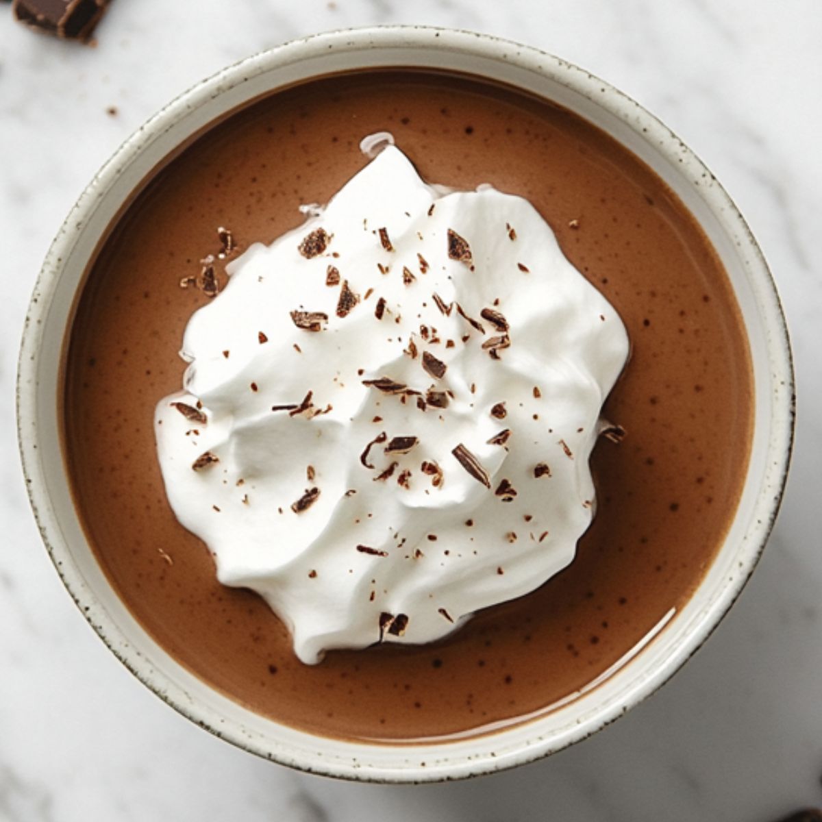 Homemade chocolate pudding recipe topped with whipped cream on a white marble kitchen counter.