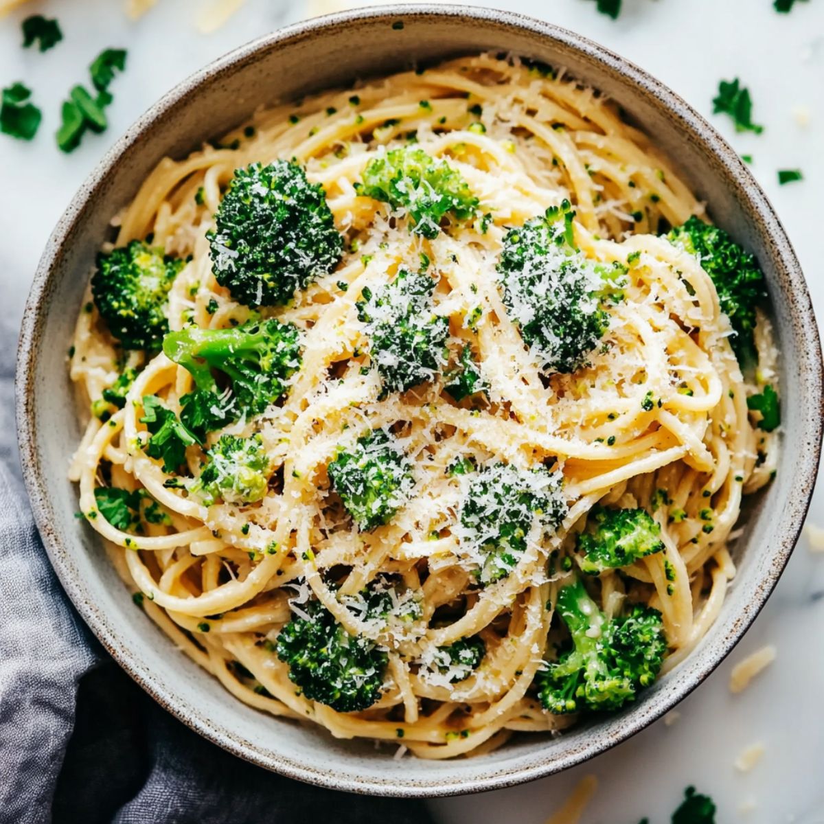 A bowl of creamy broccoli pasta, garnished with Parmesan cheese and parsley, placed on a white marble kitchen counter.