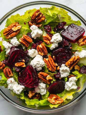 Overhead view of a colorful goat cheese salad recipe with greens, cheese, pecans, cranberries, and beets in a glass bowl on a marble counter.