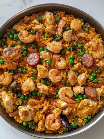 A top-view of freshly made paella recipe served in a glass pan with shrimp, mussels, chicken, and peas on white marble.