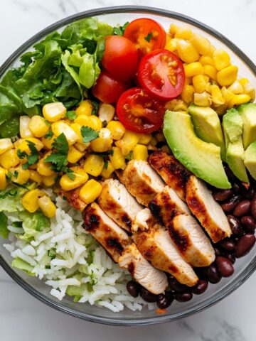Overhead view of a colorful homemade burrito bowl recipe on white marble with fresh toppings and lime.
