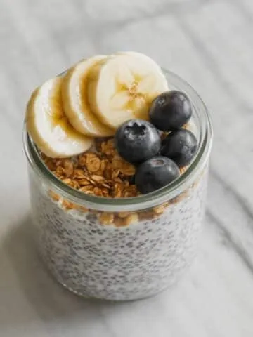 Overhead view of chia pudding recipe in a glass jar topped with fruit and granola on a white marble kitchen counter