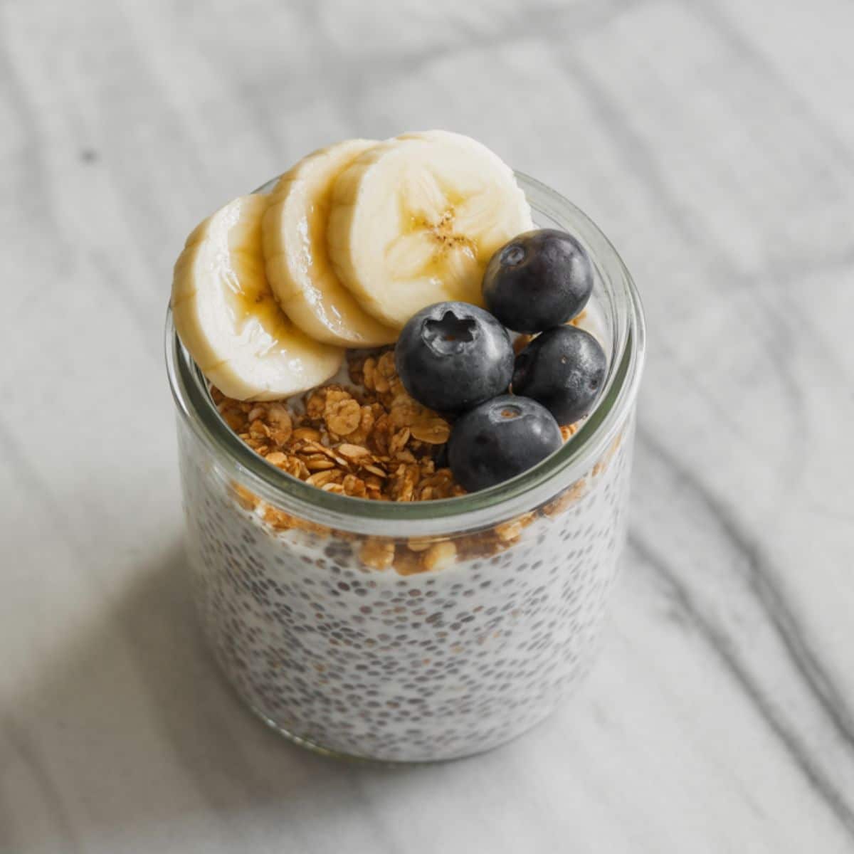 Overhead view of chia pudding recipe in a glass jar topped with fruit and granola on a white marble kitchen counter