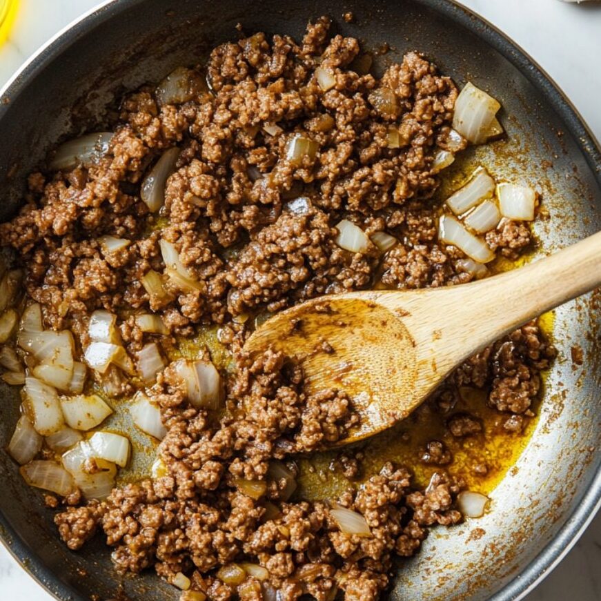 Ground beef, onions, and garlic being sautéed in a pan for cheeseburger bomb filling.