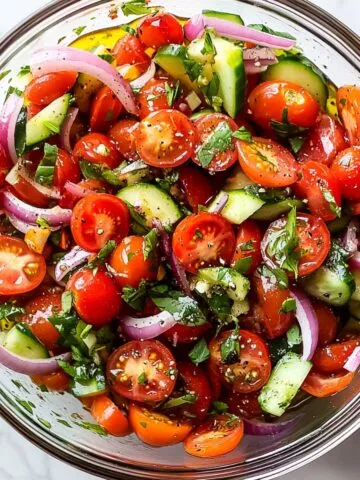Homemade tomato salad recipe with cherry tomatoes, onions, cucumbers, and fresh herbs in a glass bowl
