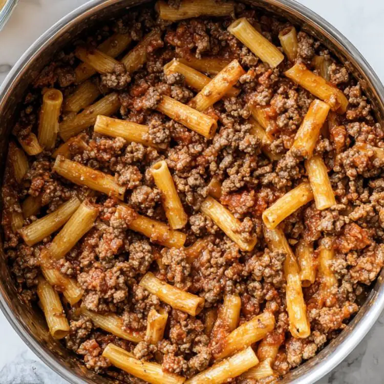 Browning ground beef and sausage next to cooked pasta and garlic on a white kitchen surface

