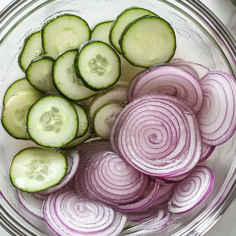 Thinly sliced red onions soaking in water and fresh cucumber slices on the counter

