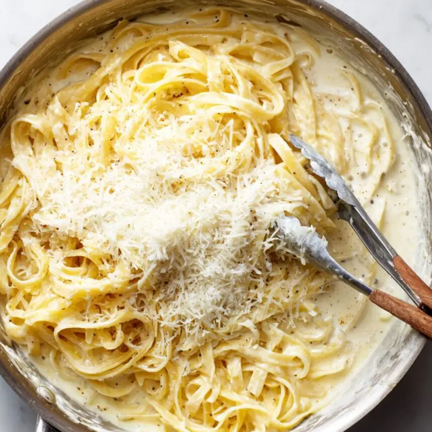 Parmesan cheese being added to Alfredo sauce with fettuccine in a pan

