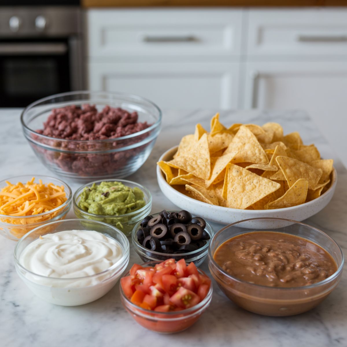 Ingredients for beef nachos recipe including ground beef, cheese, chips, and toppings on a marble kitchen counter

