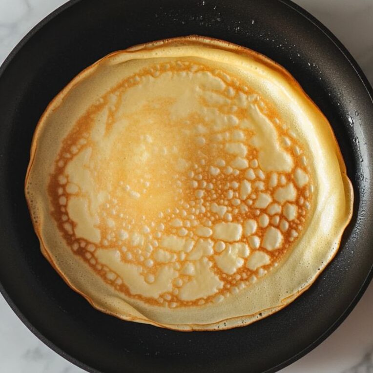 Thin crepe cooking in a non-stick pan with set edges.