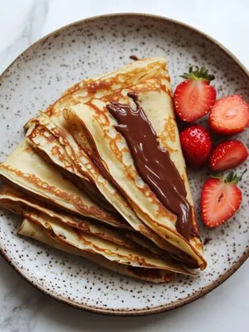 Stack of homemade Crepes Nutella recipe with strawberries on white marble counter.