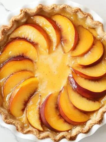 Overhead view of a freshly baked peach custard pie with golden crust and juicy peaches in a white pie dish on a marble kitchen counter.