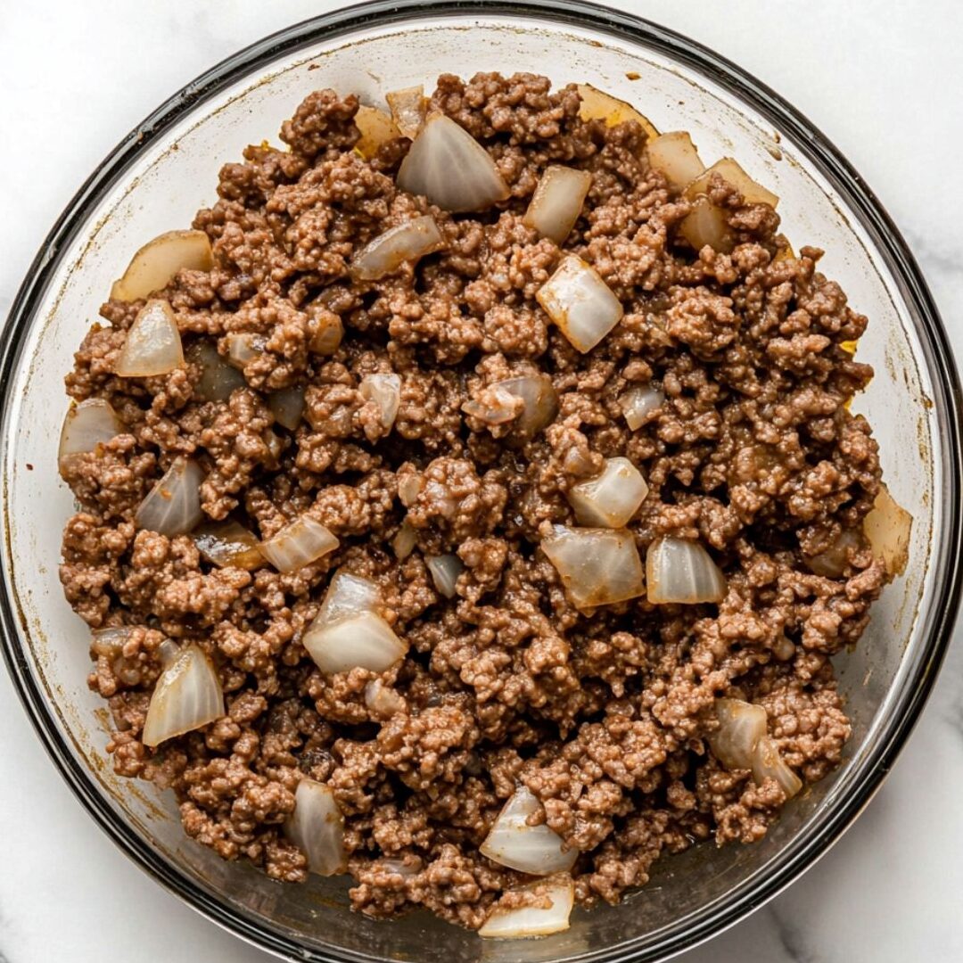 Seasoned ground beef cooking with onions and garlic in a glass skillet
