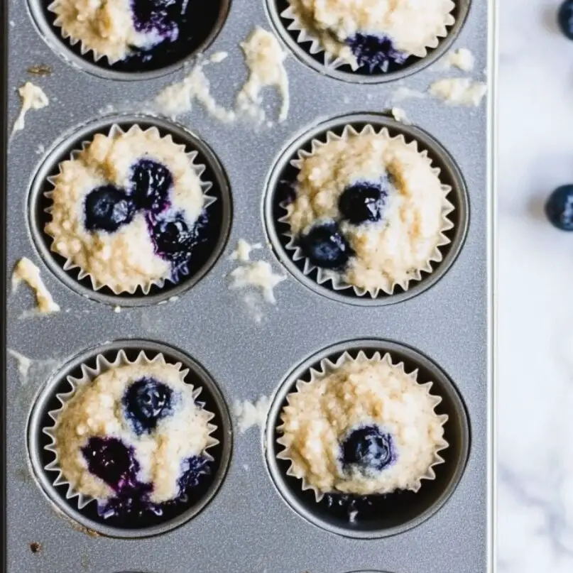 Muffin tin with unbaked blueberry muffin batter on a white marble counter