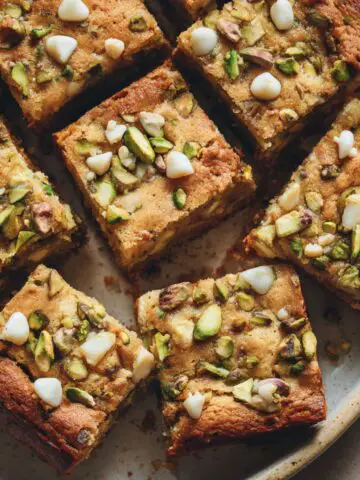 Tray of sliced pistachio blondies recipe on a white kitchen counter with chopped pistachios and white chocolate