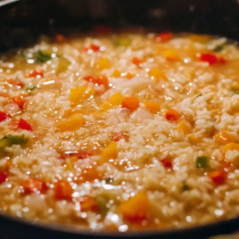 Close-up of jasmine rice cooking with onions and bell peppers in broth on a stovetop.