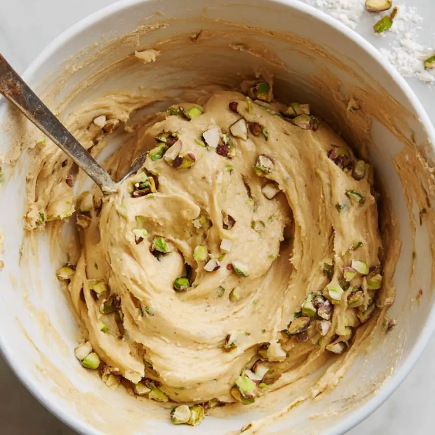 Blondie batter in a bowl with pistachios and white chocolate chips on a white marble counter