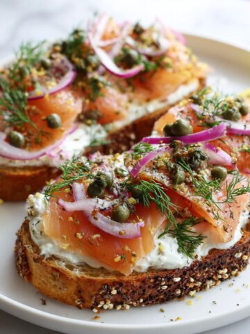 Overhead shot of smoked salmon toast recipe with cream cheese, dill, capers, and lemon on white plate.