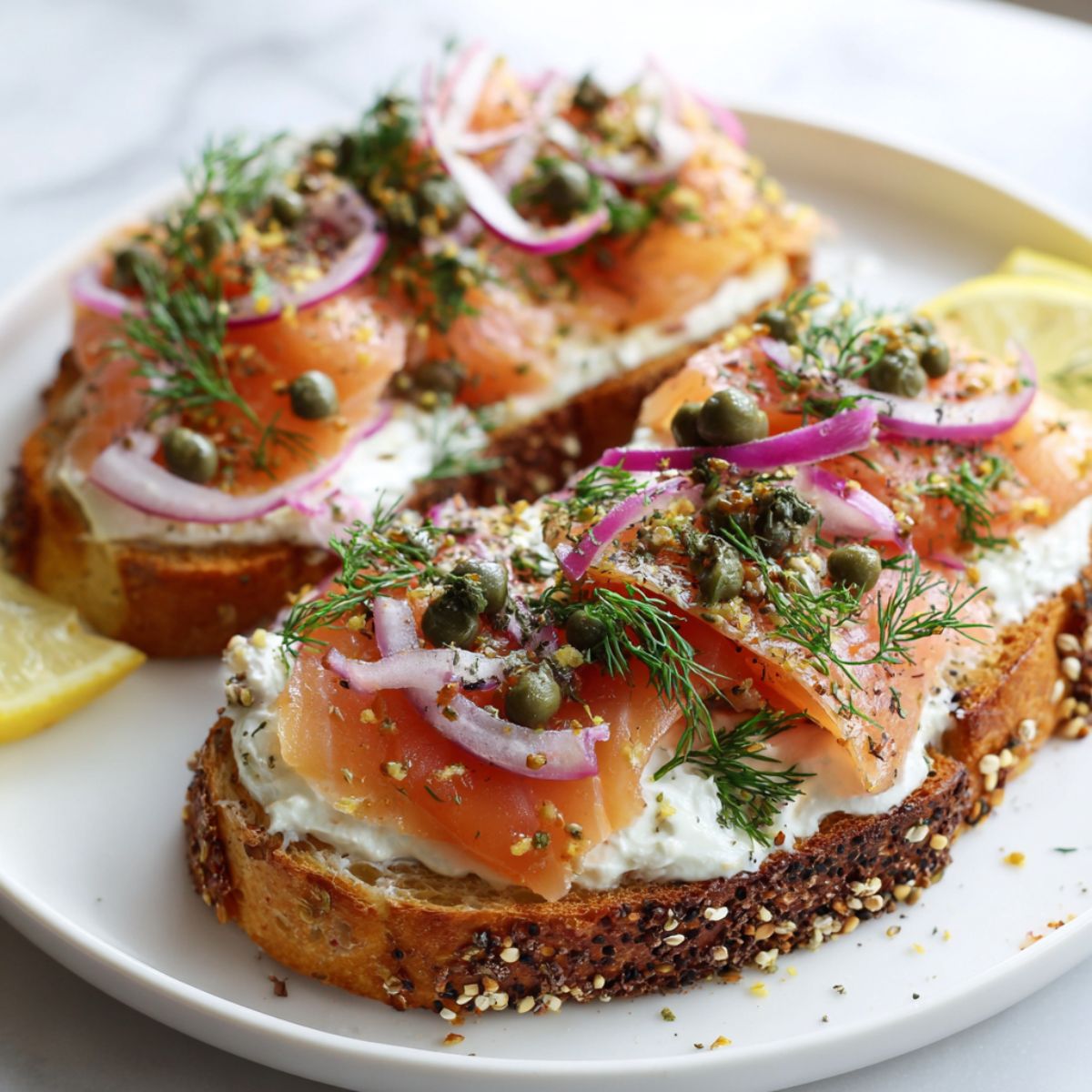 Overhead shot of smoked salmon toast recipe with cream cheese, dill, capers, and lemon on white plate.