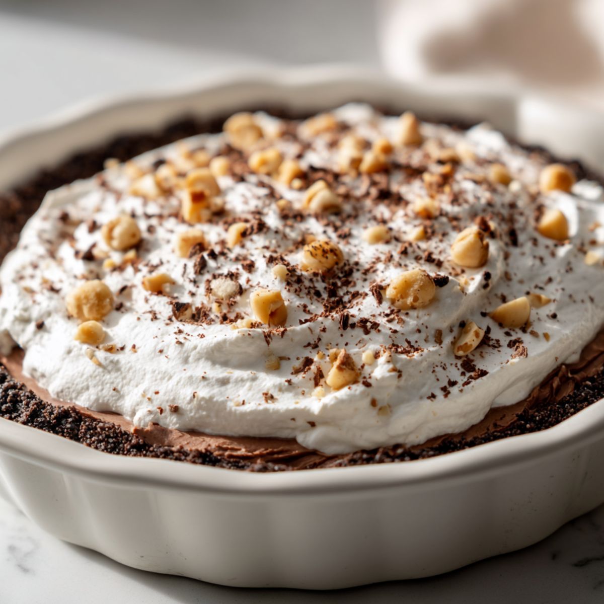 Homemade Nutella pie recipe topped with whipped cream, chocolate shavings, and hazelnuts on white counter.

