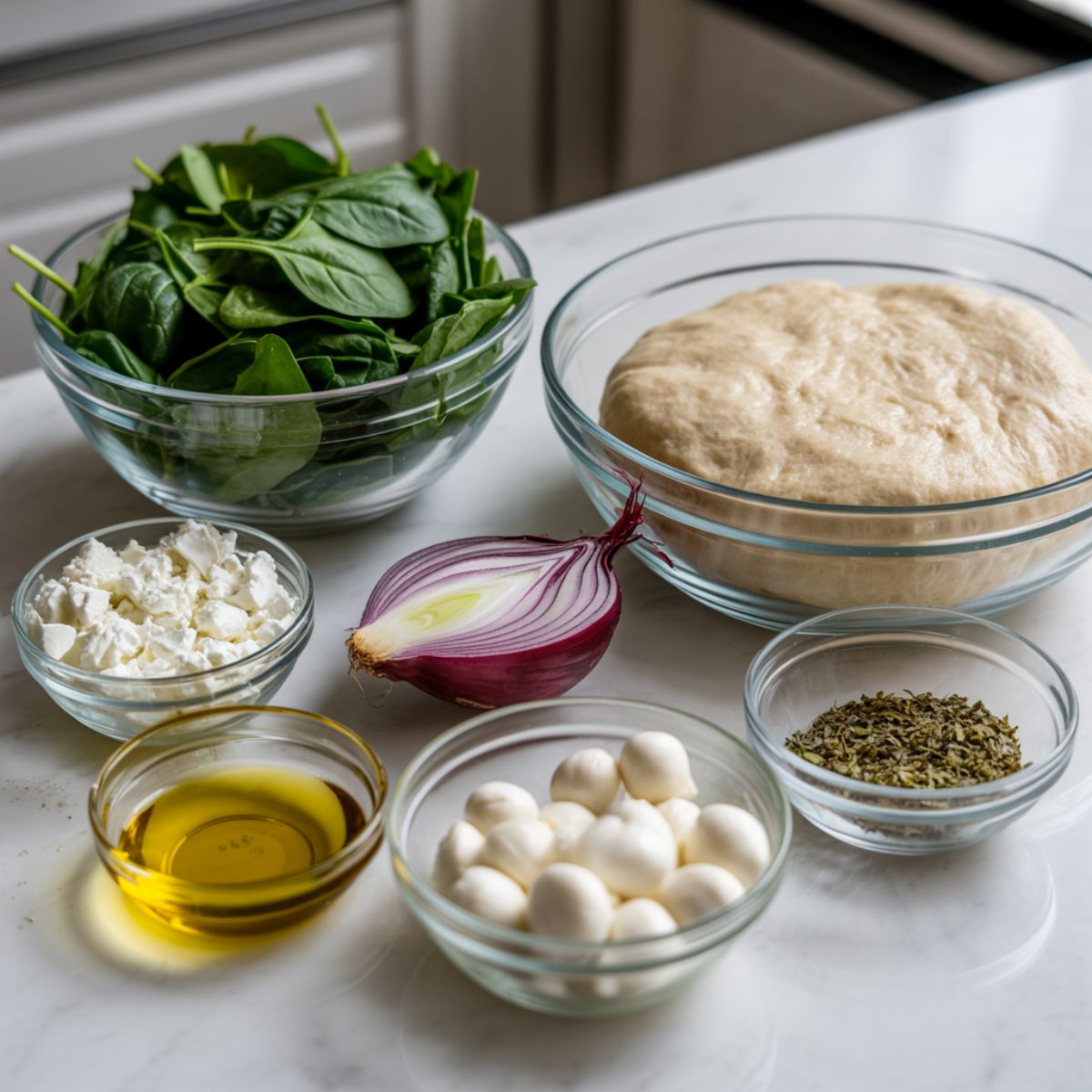 Flatlay of fresh spinach, feta, garlic, red onion, and pizza dough on a white kitchen surface