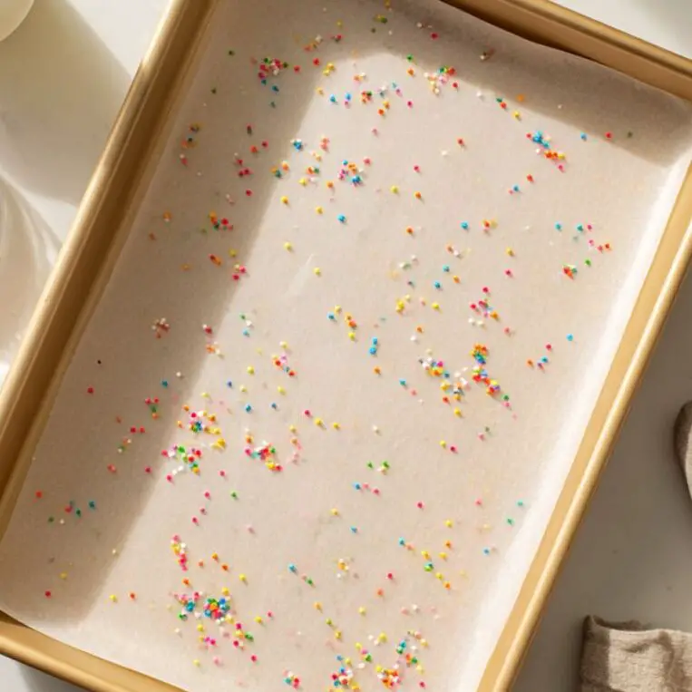 Lined baking pan with parchment paper and a bowl of sprinkles on a kitchen counter

