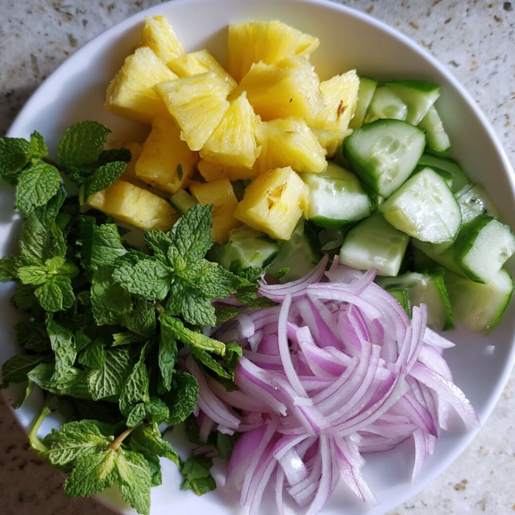 Freshly cut pineapple, cucumber, and herbs prepared on a white surface

