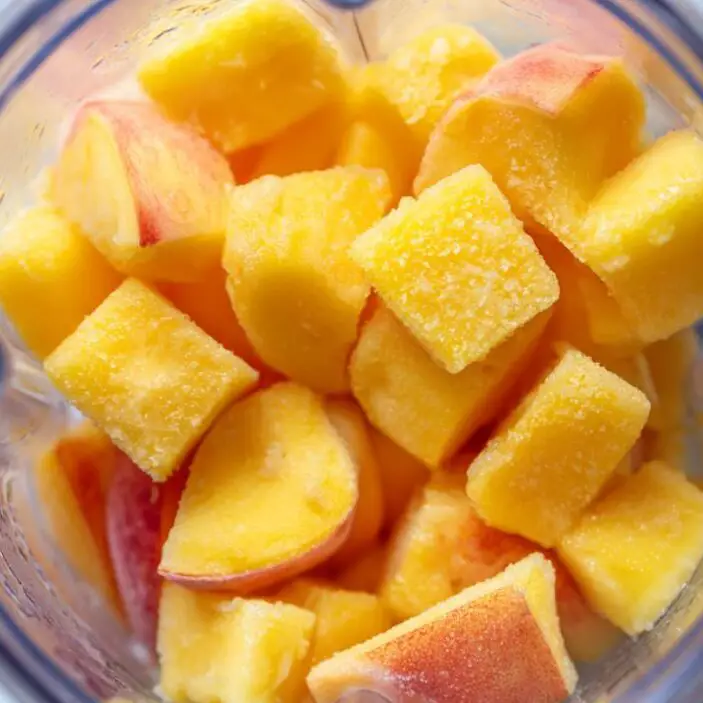 Blender with mango chunks and peach slices added, ready to blend