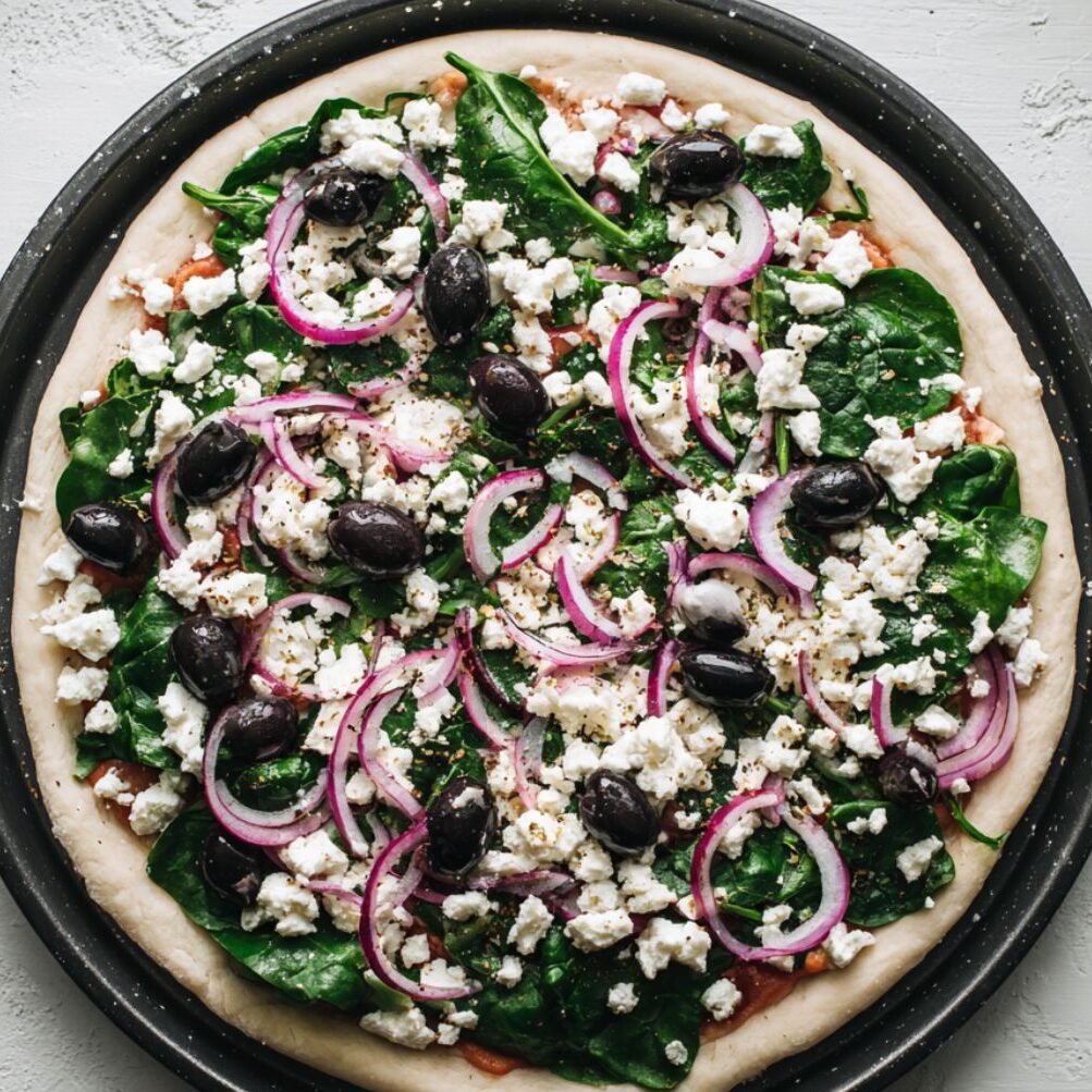 Unbaked pizza layered with spinach, red onion, feta, and cheese