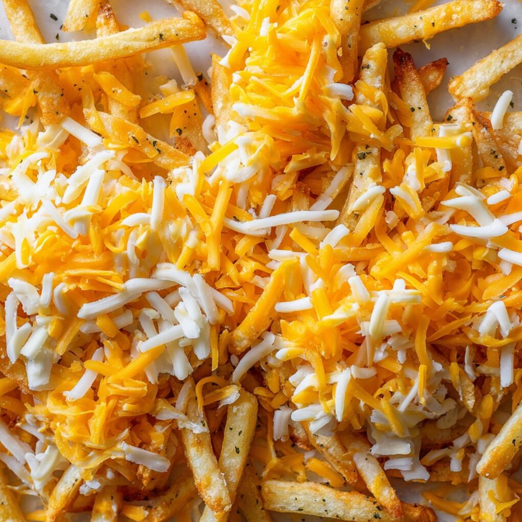 Fries being layered with shredded cheddar and nacho cheese before baking.
