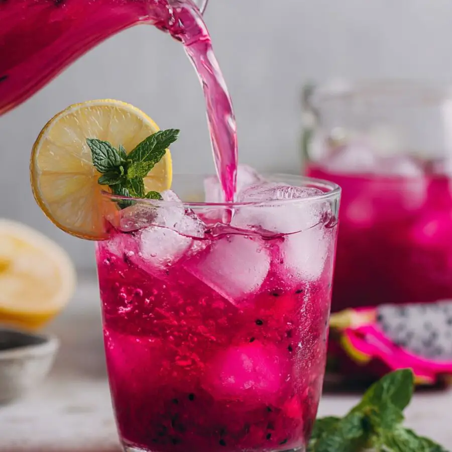 A glass of dragon fruit lemonade garnished with mint and lemon beside a pitcher.