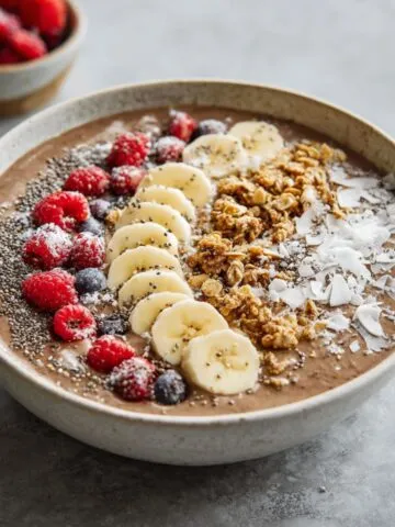 Homemade chocolate banana smoothie bowl recipe with granola, sliced bananas, chia seeds, coconut flakes, and fresh berries on a white kitchen counter, photographed from above.