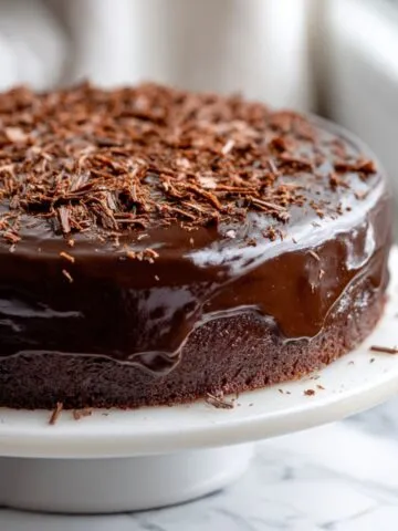 Overhead shot of a homemade chocolate truffle cake recipe with glossy ganache and chocolate shavings on a white kitchen counter.