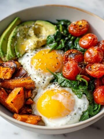 Homemade savory breakfast bowl recipe with eggs, avocado, spinach, sweet potatoes, and cherry tomatoes on a white kitchen counter, photographed from above.