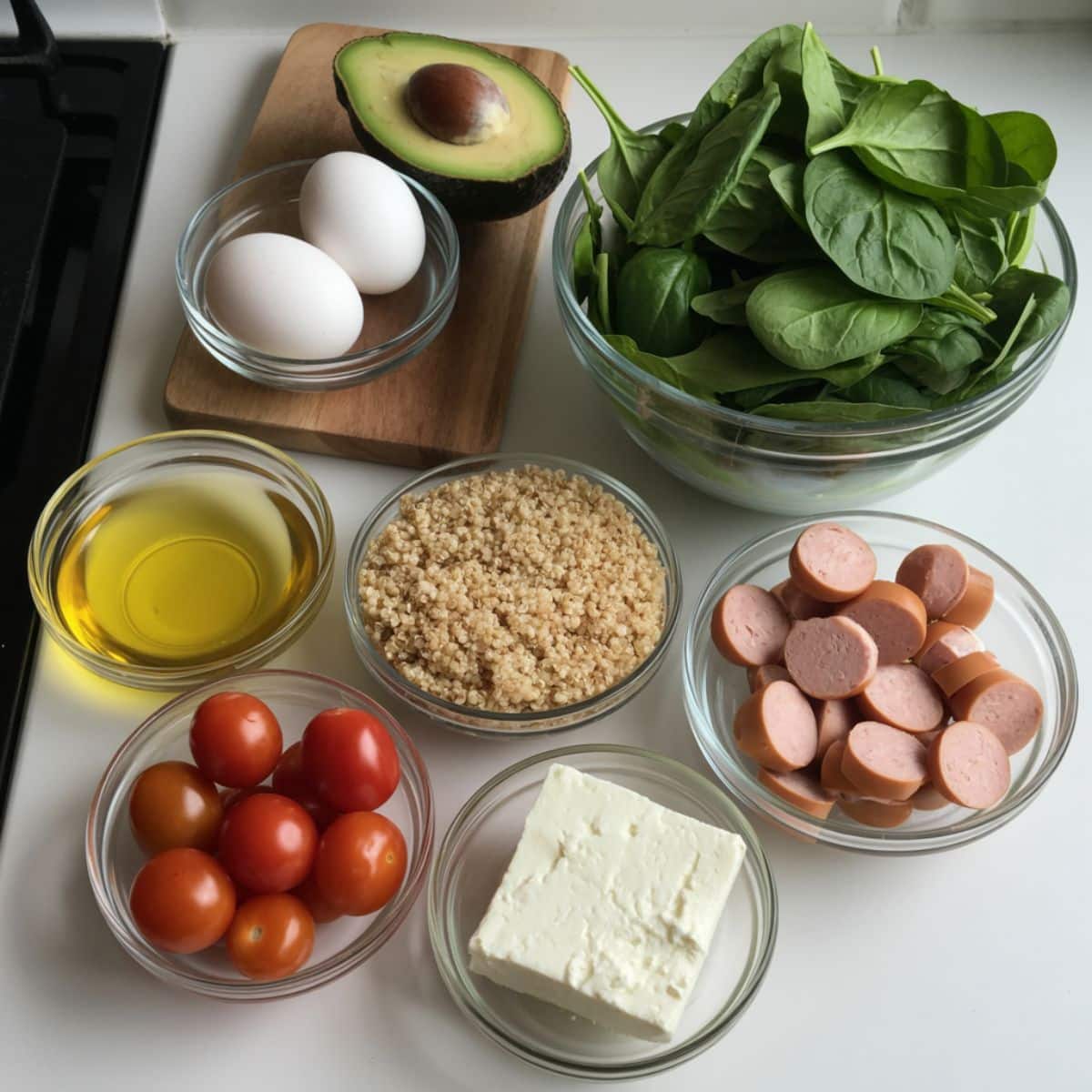 Overhead view of raw ingredients for a savory breakfast bowl including eggs, avocado, spinach, cherry tomatoes, quinoa, and sausage on a white kitchen counter.