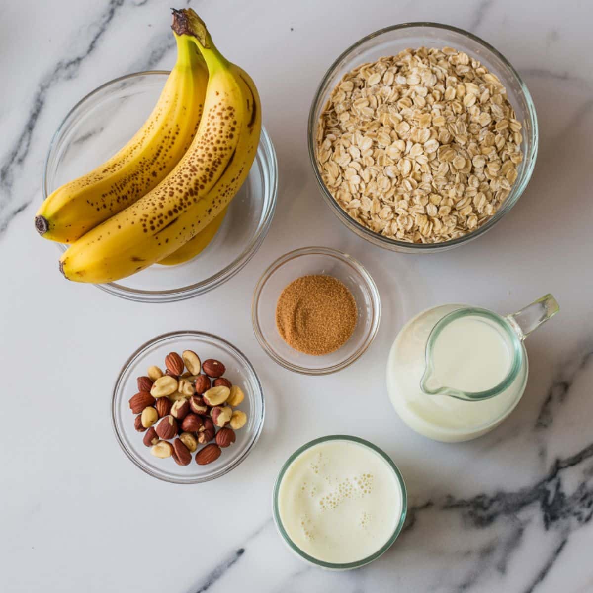 Overhead view of ingredients for banana nut oatmeal: bananas, oats, nuts, honey, and milk on a white kitchen counter.