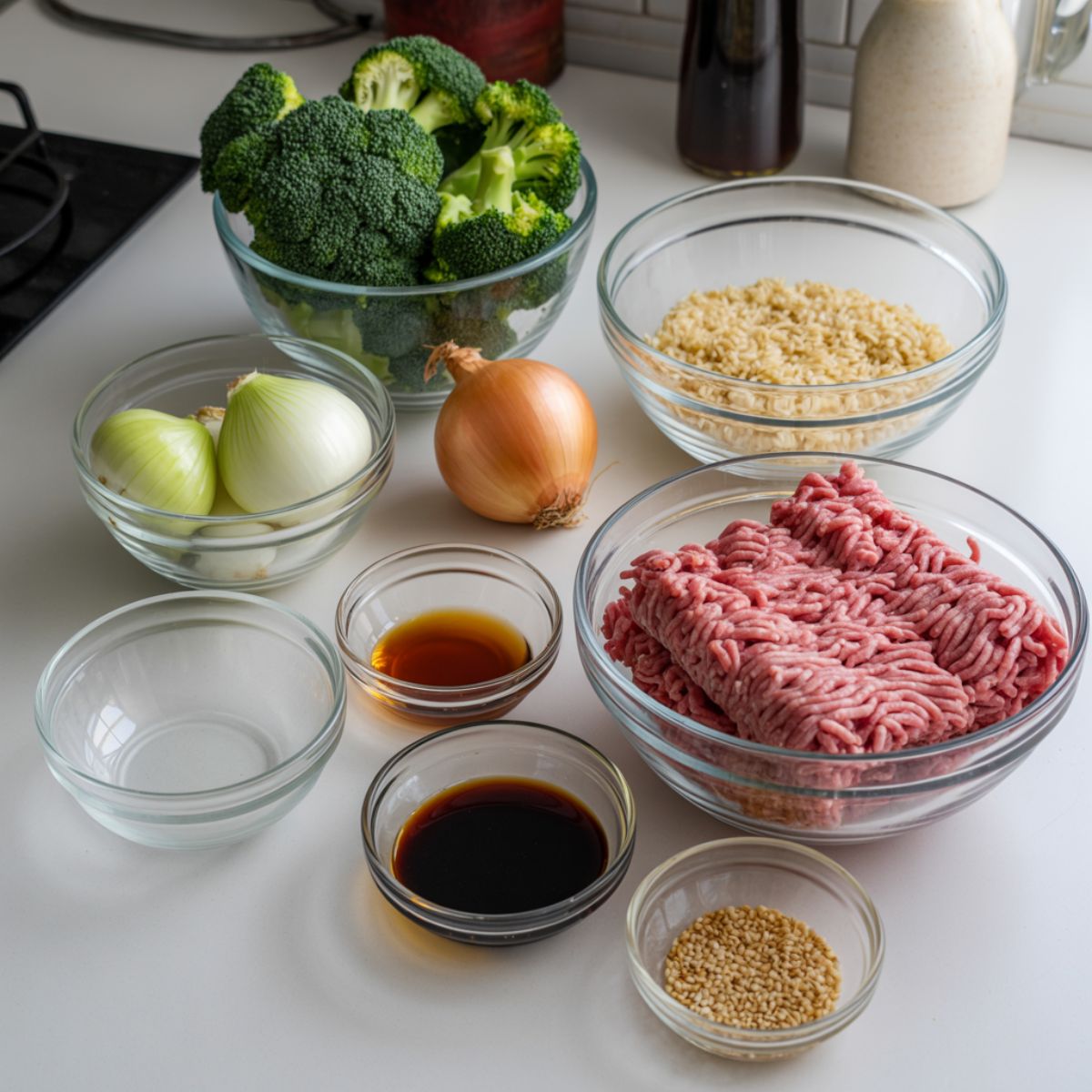 Overhead view of fresh ingredients for a beef bowl, including ground beef, rice, broccoli, onion, garlic, and sauces on a white kitchen counter.