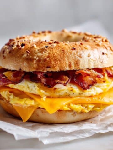 Homemade breakfast bagel sandwich recipe with egg, melted cheddar cheese, and bacon on a white kitchen counter photographed from above.