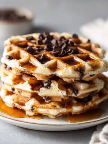 Overhead shot of homemade chocolate chip waffles recipe stacked on a white plate with melted chocolate chips and maple syrup, sitting on a white kitchen counter.
