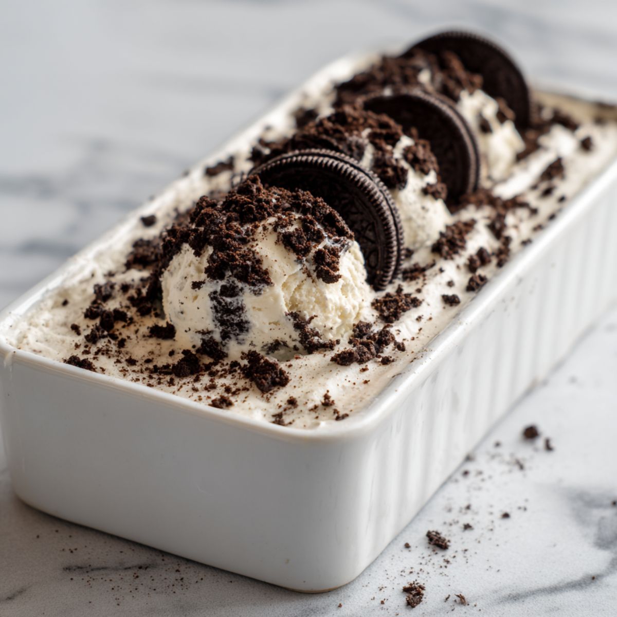 Homemade Oreo ice cream recipe in a loaf pan topped with crushed cookies on a white marble counter, photographed from above.