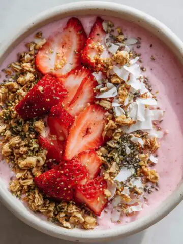 Homemade strawberry smoothie bowl recipe with fresh strawberries, granola, and coconut flakes on a white kitchen counter, overhead view.