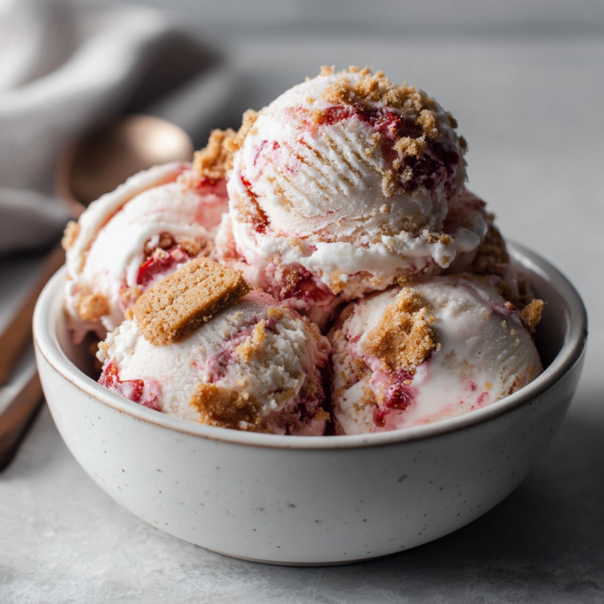 Bowl of homemade strawberry cheesecake ice cream recipe with strawberry swirl and graham cracker pieces on a white marble counter.