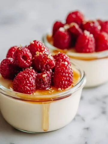 Homemade vanilla bean panna cotta recipe in glass ramekins topped with fresh raspberries and honey drizzle, photographed overhead on a white marble counter.