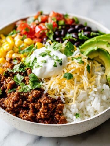 Homemade beef burrito bowl recipe with rice, beans, avocado, corn, cheese, and sour cream served on a white kitchen counter, photographed from above.