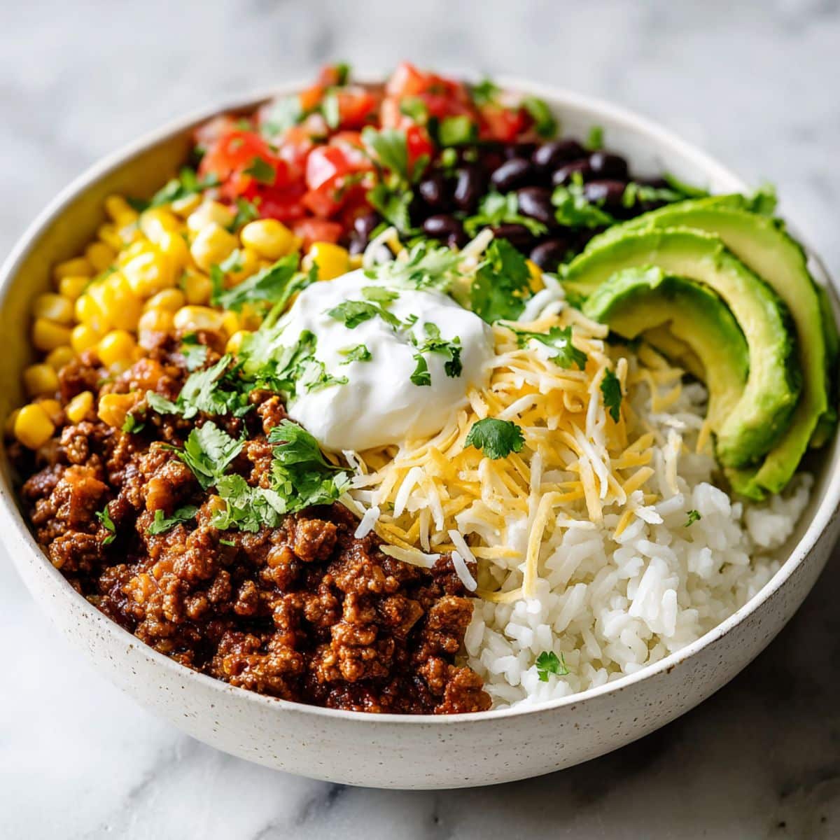 Homemade beef burrito bowl recipe with rice, beans, avocado, corn, cheese, and sour cream served on a white kitchen counter, photographed from above.