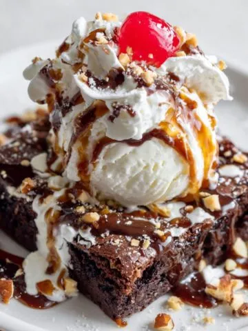 Homemade chocolate brownie sundae recipe with ice cream, fudge, caramel, whipped cream, and cherry on a white kitchen counter, photographed from above.