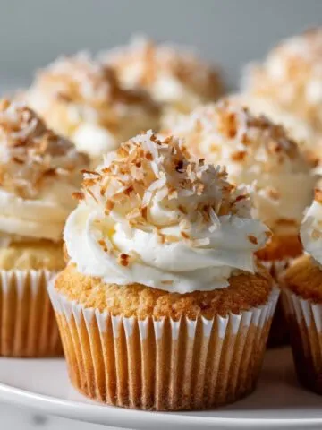 Homemade coconut cream cupcakes recipe with coconut frosting and toasted coconut flakes on a white kitchen counter, shot from above.